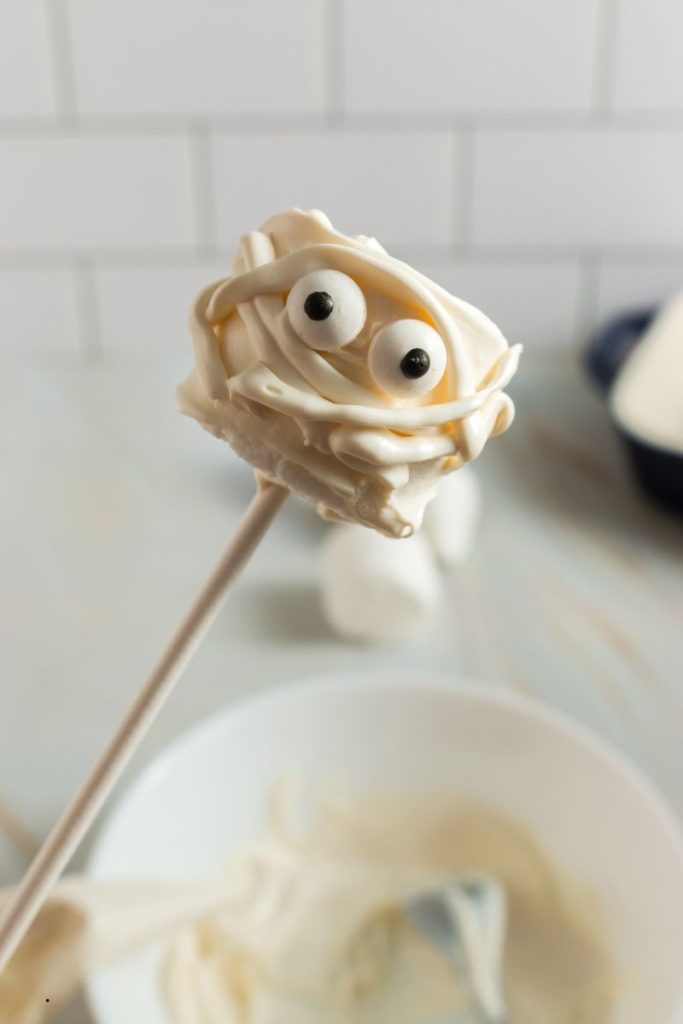 a marshmallow on a stick with two eyes and white chocolate bandages