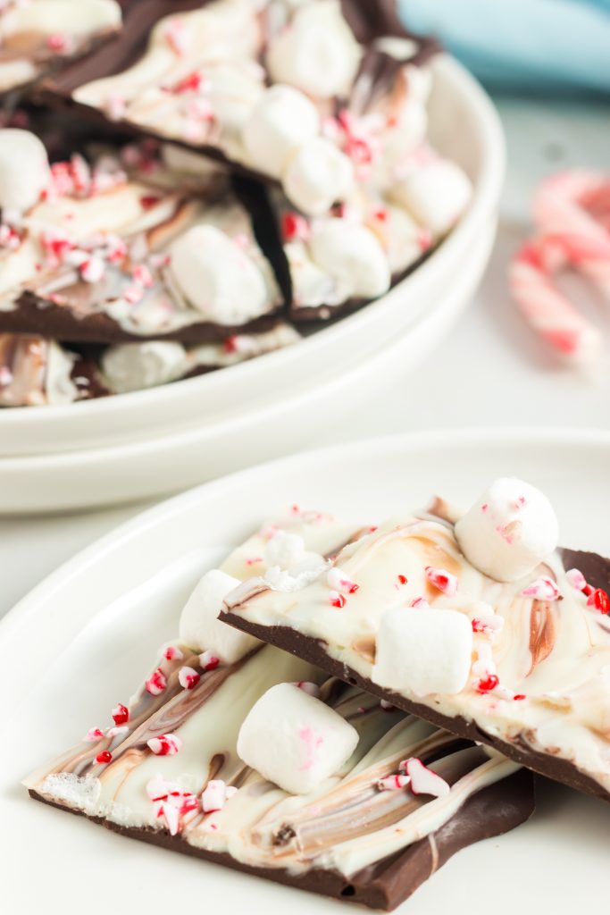 two white plates. The one in the back has a pile of brown and white chocolate bars. The plate in front has two white and brown chocolate bars with a sprinkle of marshmallows and crushed candy canes