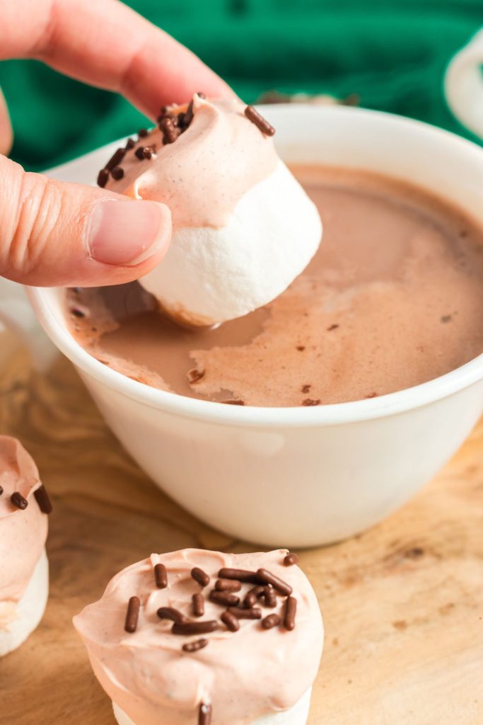 a hand holding a marshmallow over a cup of hot chocolate