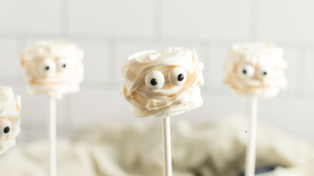 Three marshmallows mummies on individual lollipop sticks. They have eyes and white chocolate gauze around them
